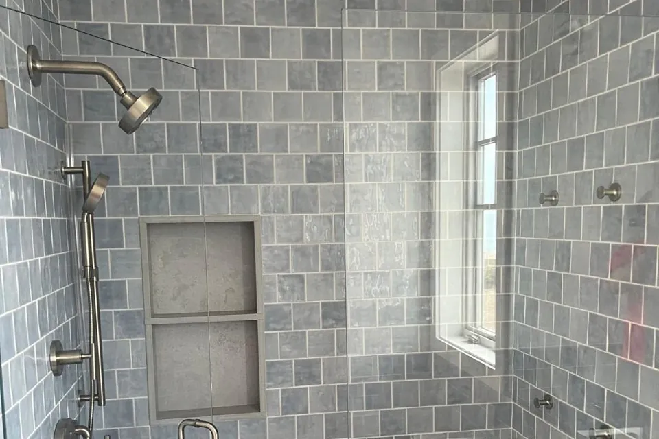 Finished bathroom shower with blue-gray square tile, recessed shelves and a glass door