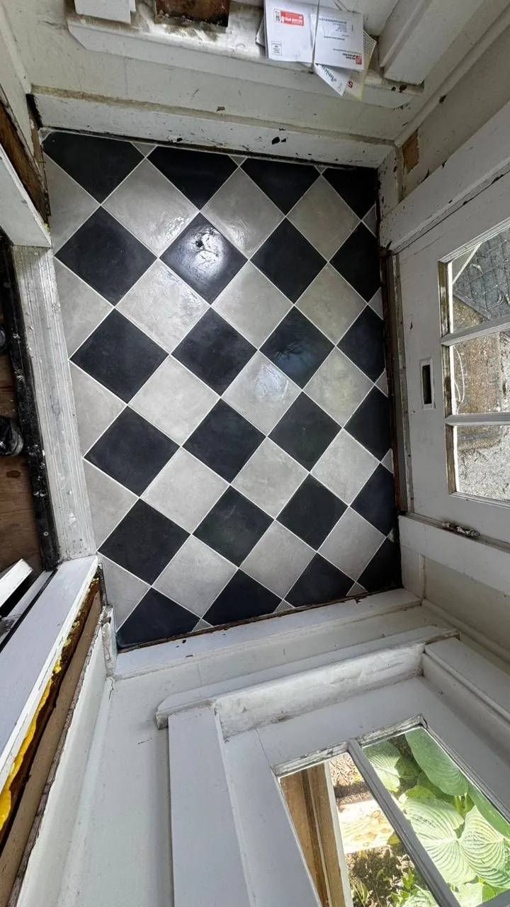 Old black and gray checkerboard tile floor in a room under demolition, with exposed framing and window trim